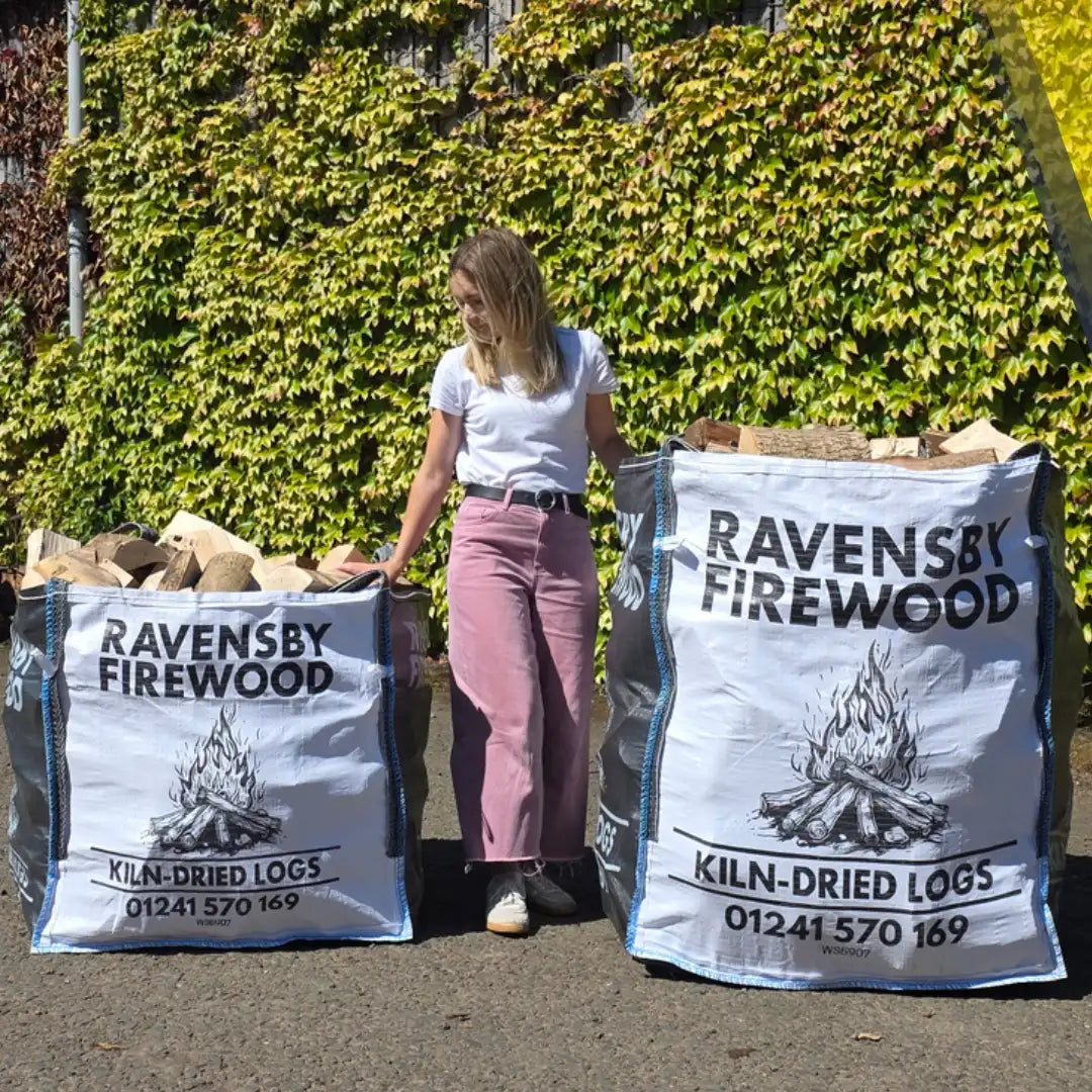 A woman stands outdoors between two large bulk bags of Ravensby Firewood kiln-dried logs, with a leafy green ivy-covered wall behind her.
