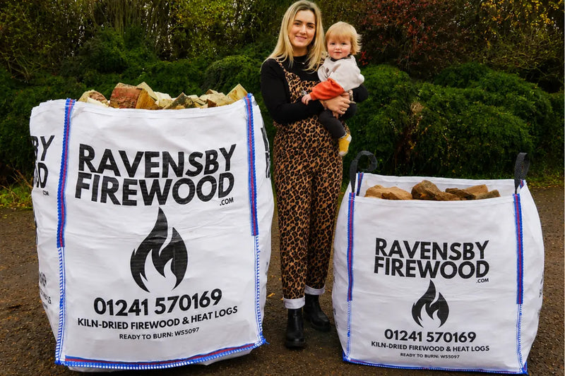 Woman holding a child in a Carnoustie garden standing next to two firewood bulk bags of kiln-dried hardwood logs with Ravensby Firewood branding.