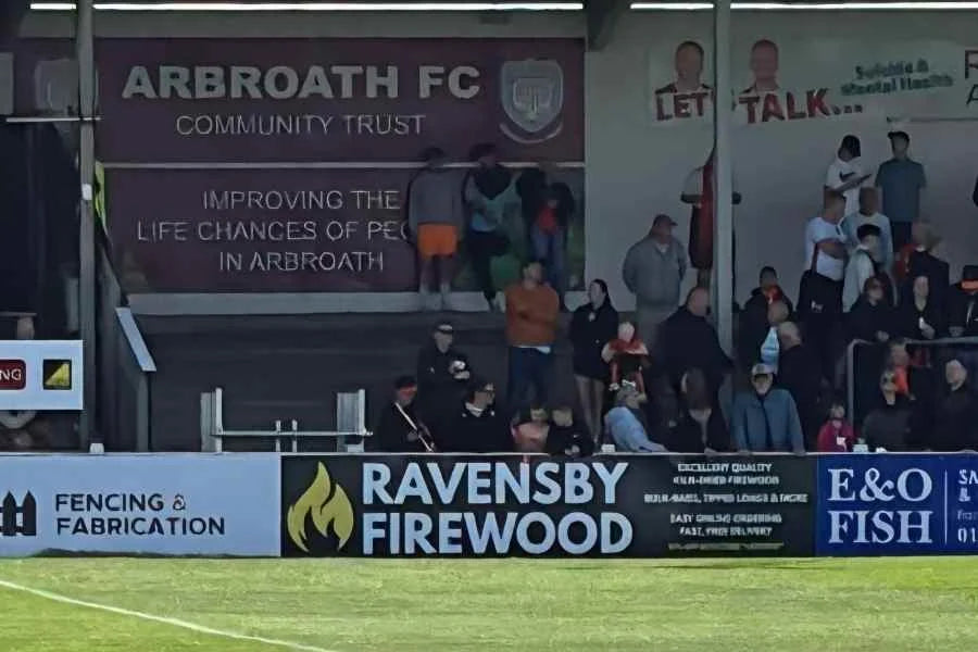 Spectators at a sports event with Arbroath FC branding and Ravensbty Firewood advertisement hoarding at side of pitch.